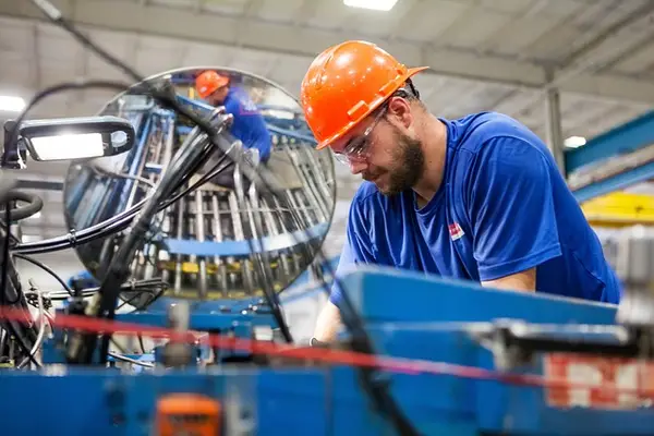 Worker in blue shirt and orange safety helmet operating machinery in industrial setting, emphasizing security guard services in manufacturing