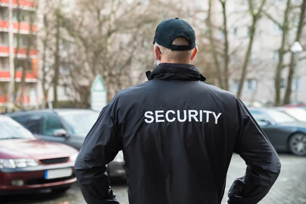 Security personnel in black jacket with 'SECURITY' monitoring parking area, ensuring safety among parked cars and trees