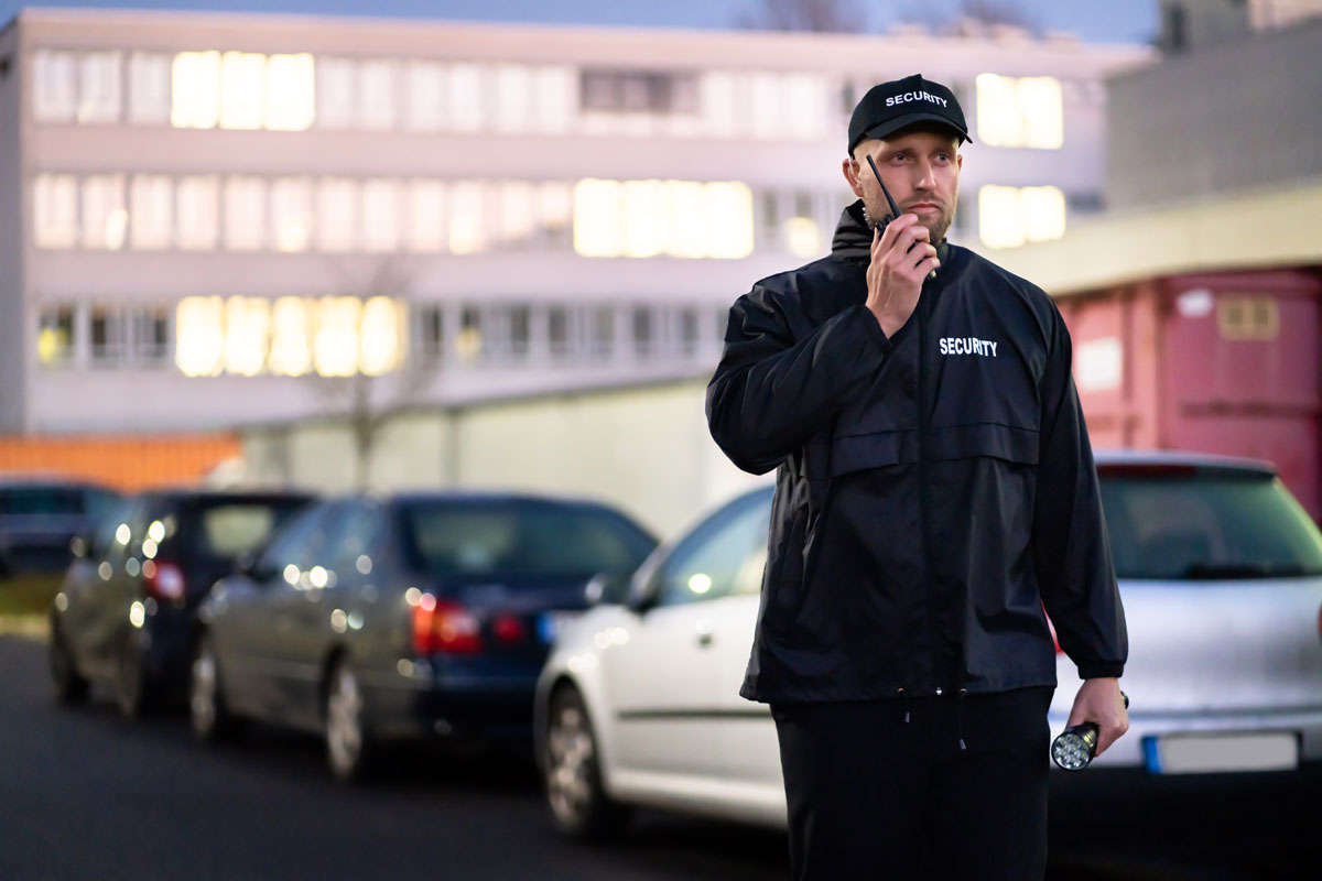 Parking security guard in black jacket with 'SECURITY' holding flashlight, monitoring vehicles in parking lot at dusk