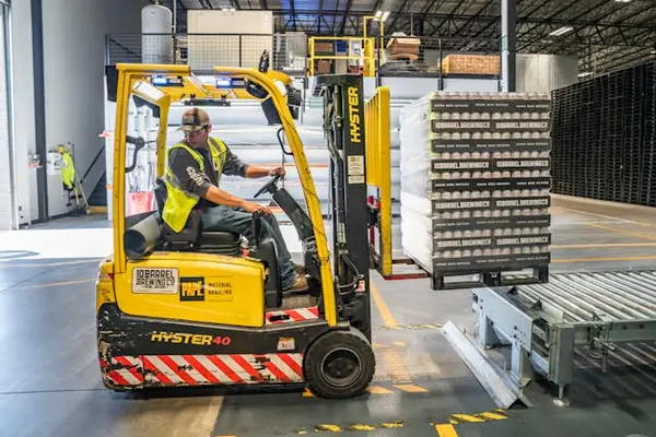 Yellow forklift operated by individual in high-visibility vest transporting goods, highlighting security guard services in warehouse environments