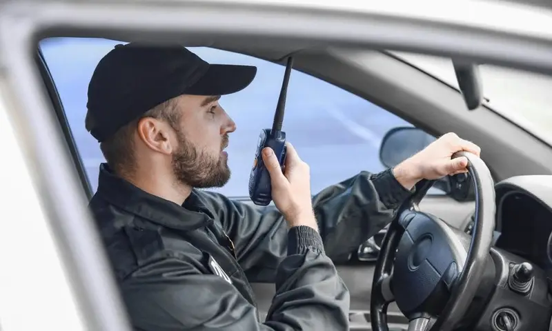 Security guard in dark uniform driving vehicle, using two-way radio to monitor and respond to mobile patrol security needs