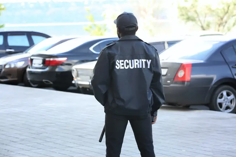 Security guard in black jacket with 'SECURITY' printed on back, monitoring parking lot with parked cars and greenery in outdoor setting